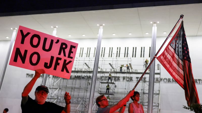 A man waves a U.S. national flag while another holds up a poster, as workers prepare scaffolding preparing ahead of removing lettering from the facade of the John F. Kennedy Memorial Center for the Performing Arts, following a federal judge’s order to remove U.S. President Donald Trump's name from the institution, in Washington, D.C., U.S., June 13, 2026.