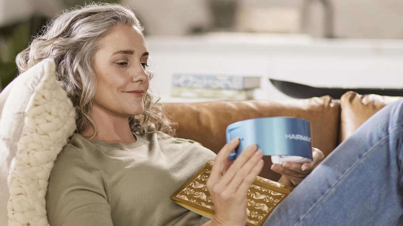 A woman with silver hair relaxes on a brown leather couch, holding the HairMax LaserBand 82 ComfortFlex device and examining it.