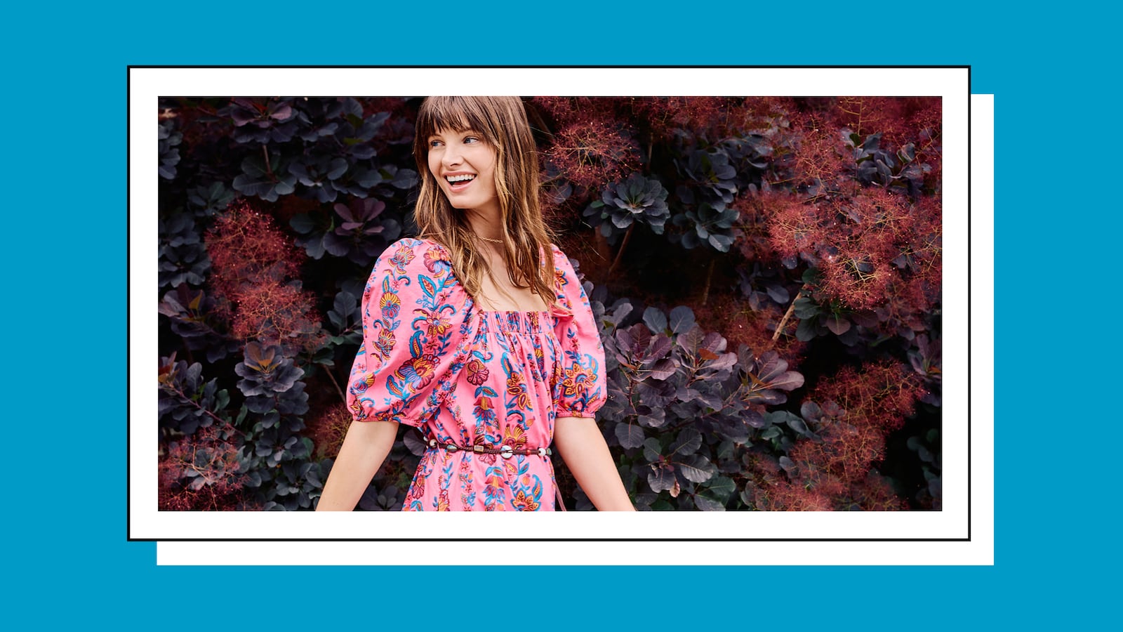A woman wearing a fuchsia dress with a floral design standing in front of a wall of flowers.