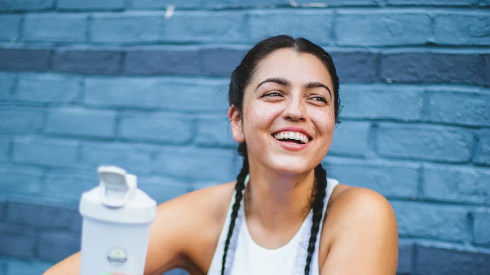 Smiling woman with braided hair holding a white shaker bottle in front of a blue brick wall.