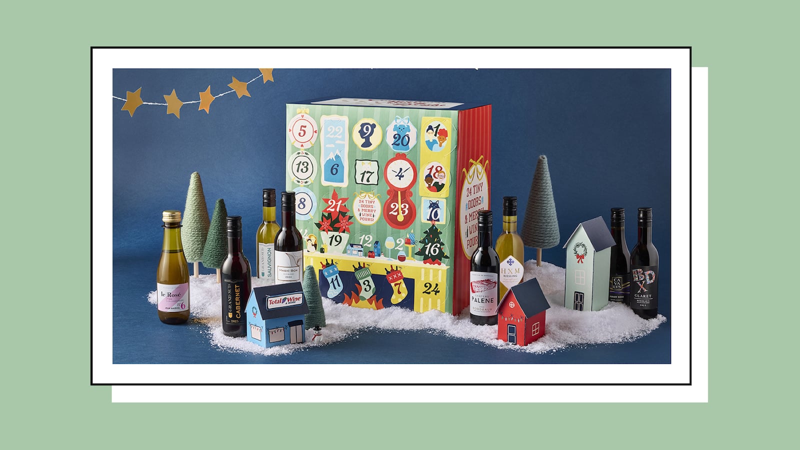 A green and red wine advent calendar with numbered doors 1 through 24, surrounded by mini wine bottles, yarn Christmas trees, and small paper houses on a snow-like display."