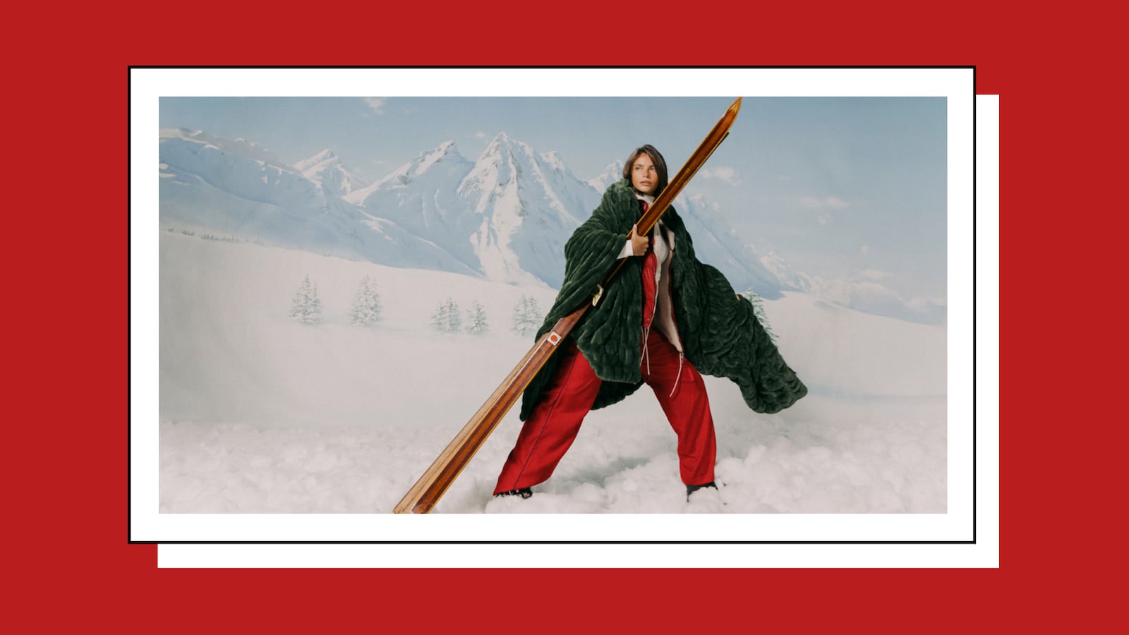 A woman poses confidently in a snowy mountain scene, wearing a deep green oversized fur coat and red pants while holding vintage wooden skis. The backdrop features tall, snow-covered peaks under a pale blue sky, evoking a luxury winter fashion editorial aesthetic.
