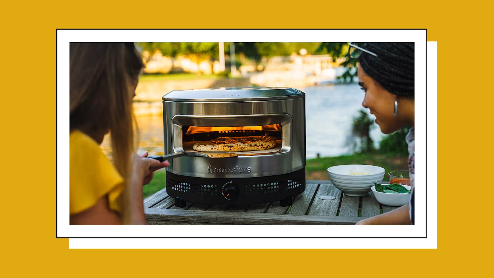 Two friends making a pizza outdoors with the Solo Stove Pi Prime pizza oven on a wooden table near a lake, stainless steel oven glowing with heat and fresh pizza baking inside.