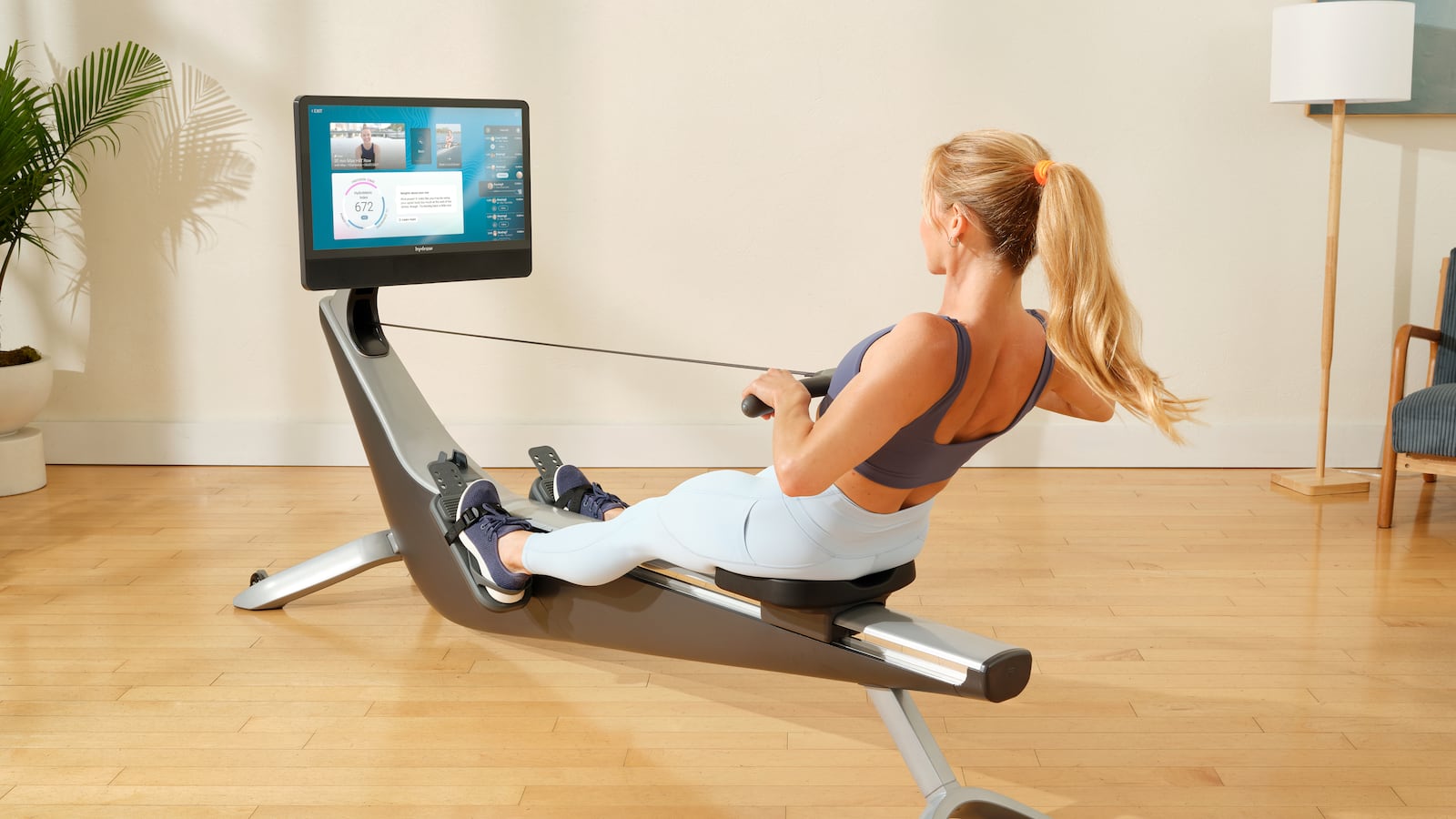 A woman in workout clothes with a blonde ponytail leans back as she works out on a Hydrow Arc Rower, which is sleek and silver with a large touchscreen. The room is light, bright, and sparse, with light hardwood floors, a floor lamp and a potted palm.