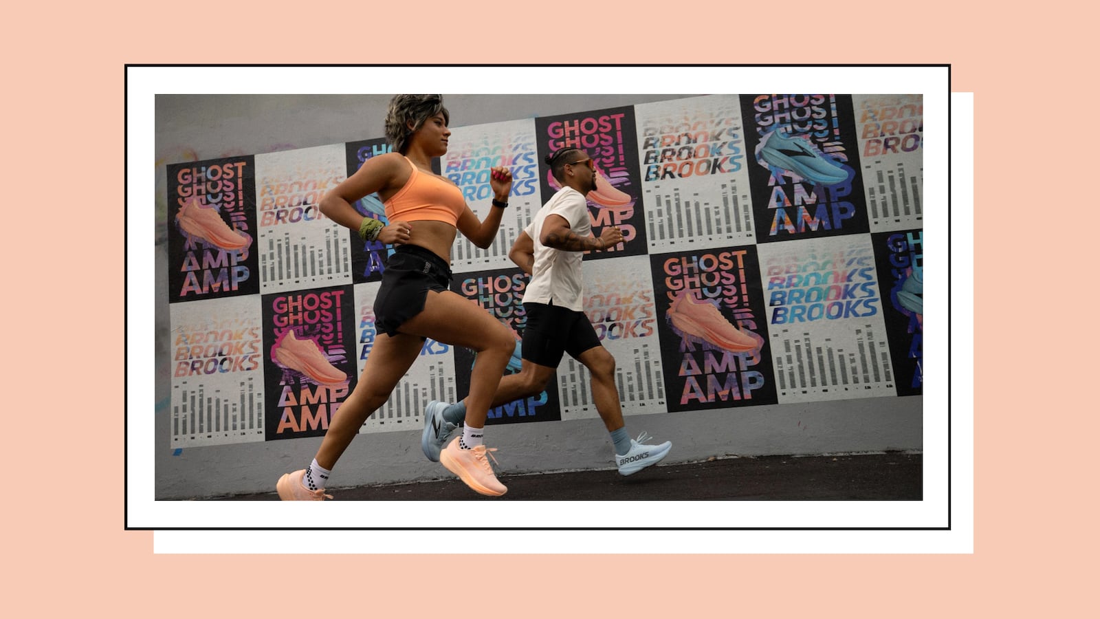 Two runners — a woman in a peach sports bra and black shorts, and a man in a white tee and black bike shorts, both in Brooks running shoes — jog past a wall covered in colorful Brooks "Ghost"/"AMP" ad posters, framed with a white and black border on a peach background.