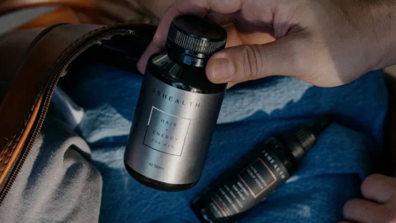 A photo of a person's hand holding up a bottle of supplements. In the background is another bottle lying on a soft surface like a bed.