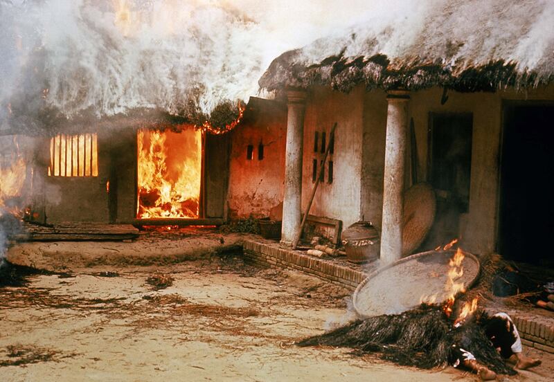 A thatched building on fire with a body in the foreground