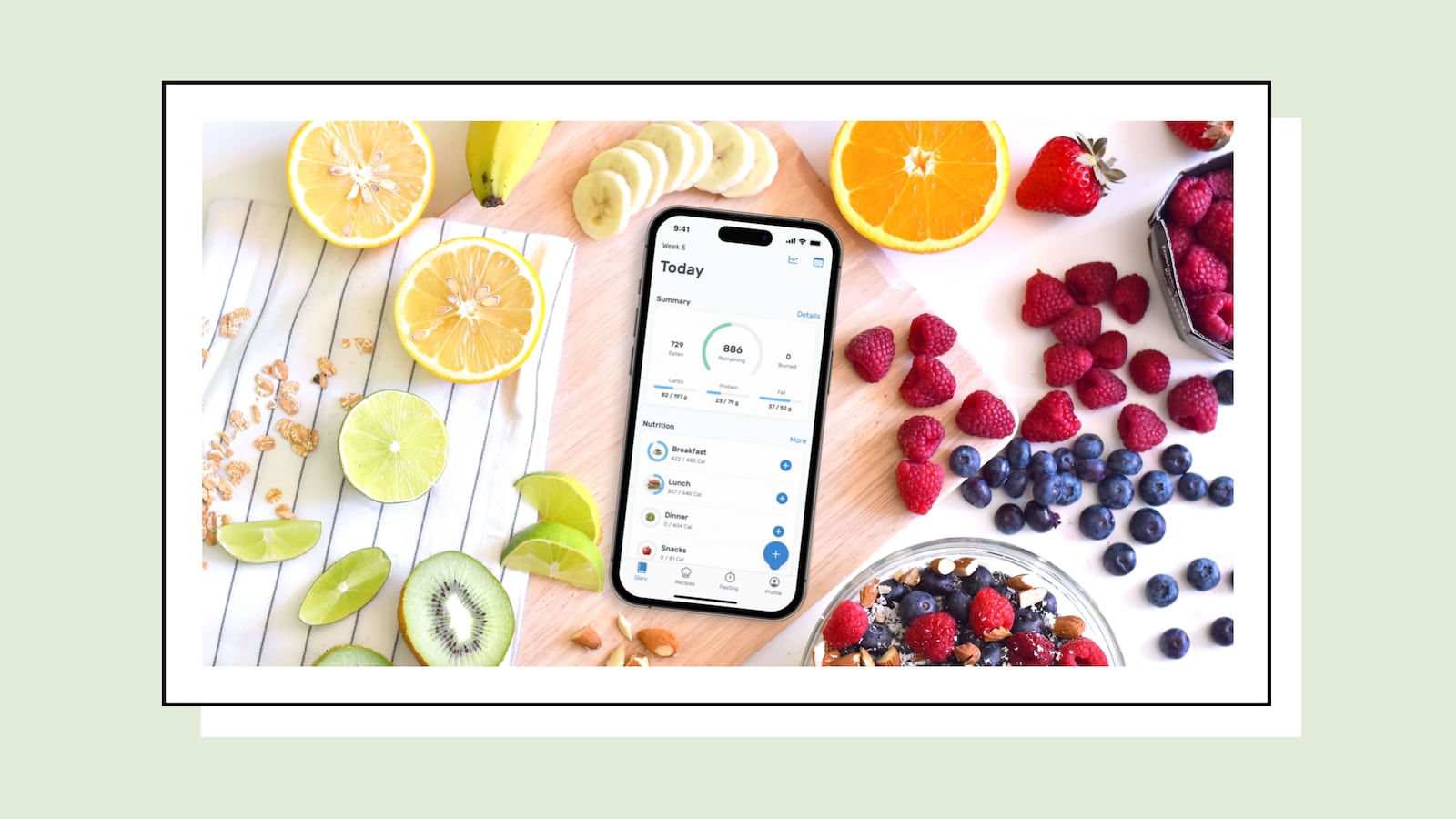 A smartphone placed on a cutting board, surrounded by an assortment of fresh fruit including strawberries, blueberries, orange slices, banana slices, and lime wedges.