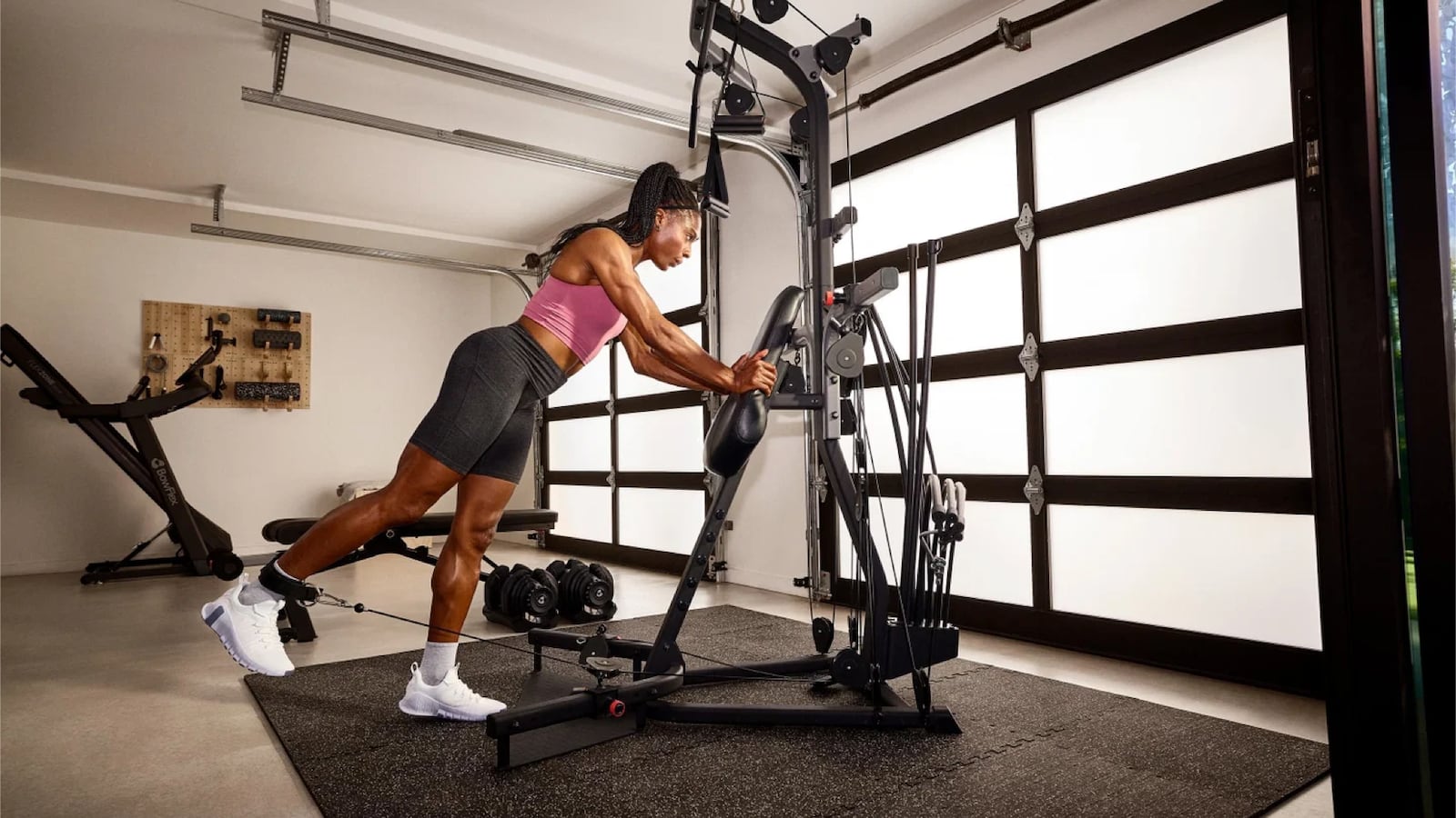Woman performing a standing cable press on the BowFlex Xtreme 2 SE home gym in a garage gym setup, with a treadmill, dumbbells, and workout bench visible in the background.