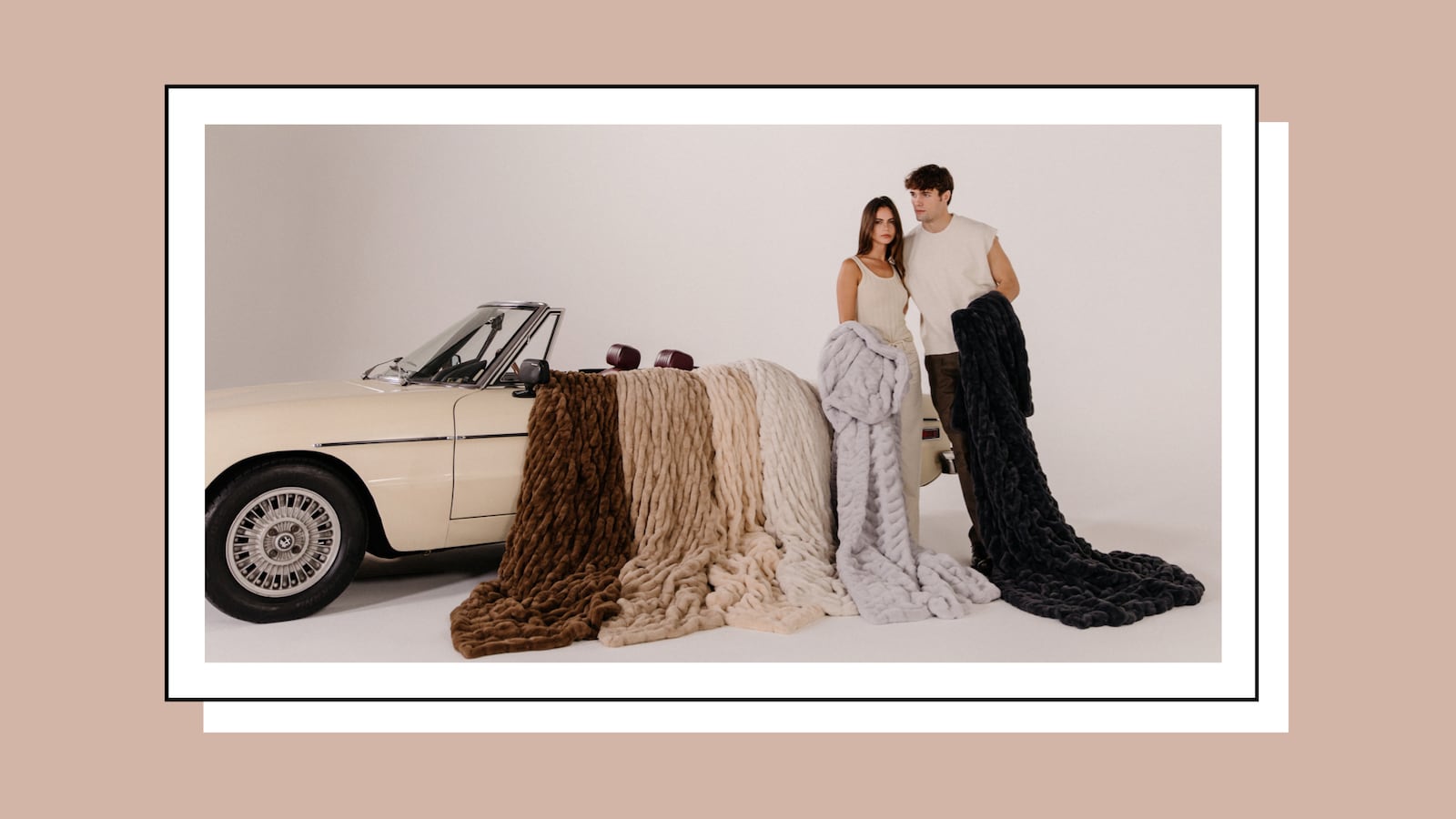 A dark-haired man and woman posing next to a convertible car. The man is holding a black blanket and the woman is holding a grey blanket. On the car there are brown, tan, peach, and white blankets laid out.
