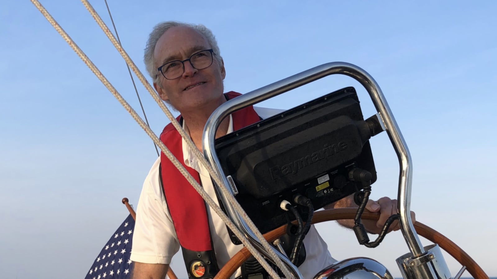 Man at the helm of a sail boat with the American flag behind him.
