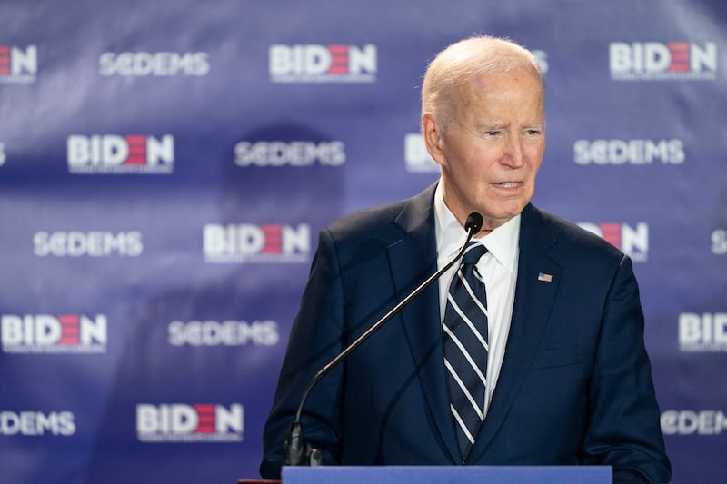 Former President Joe Biden speaking at a podium in front of a banner with 'BIDEN' and 'S-C-DEMS' written all over it.