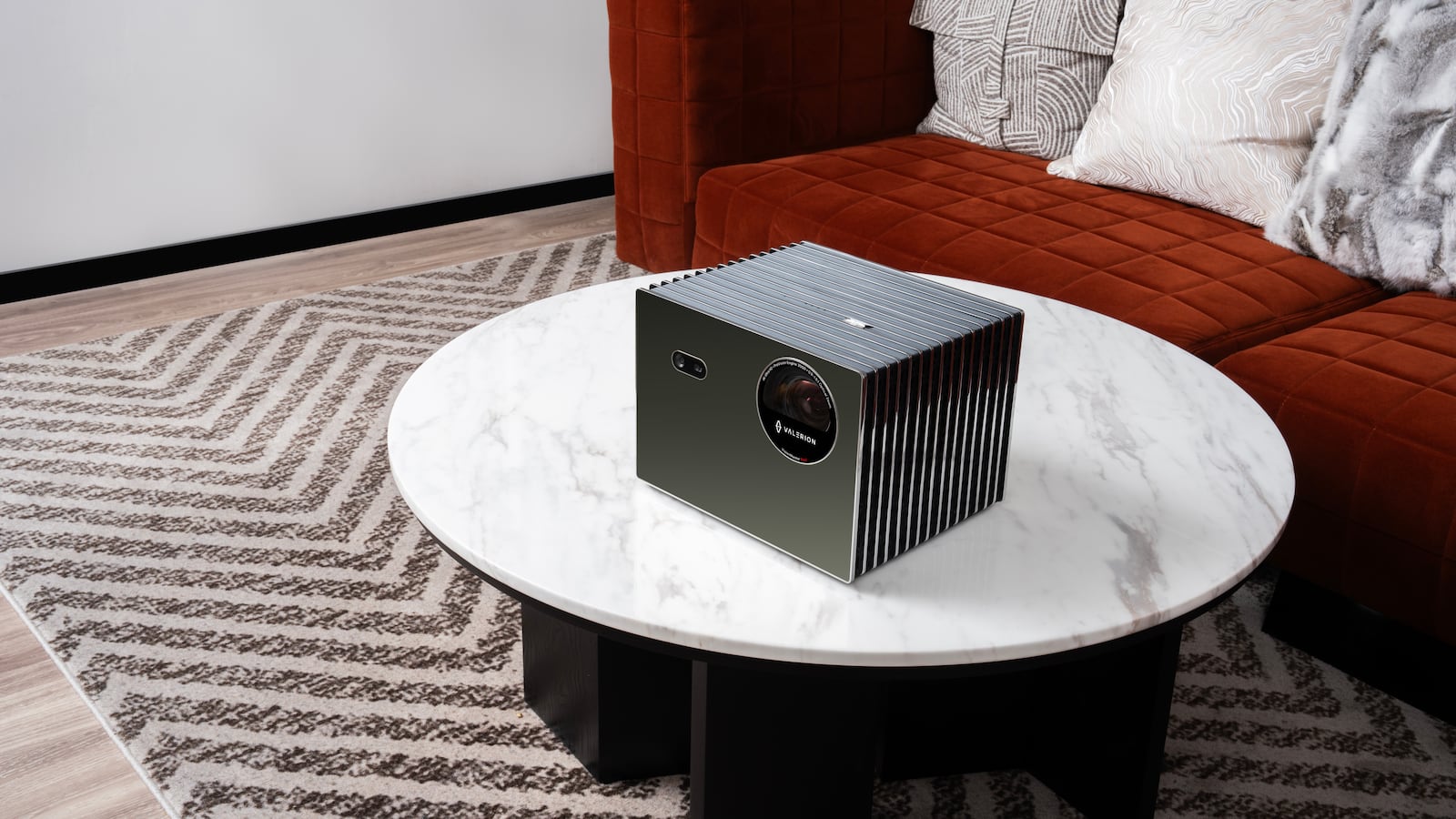 The Valerion VisionMaster Pro 2, a sleek, square projector, sits on a marble coffee table. A worn chevron rug covers gray wooden flooring. The couch is a rust red velvet with patterned white-on-white throw pillows.