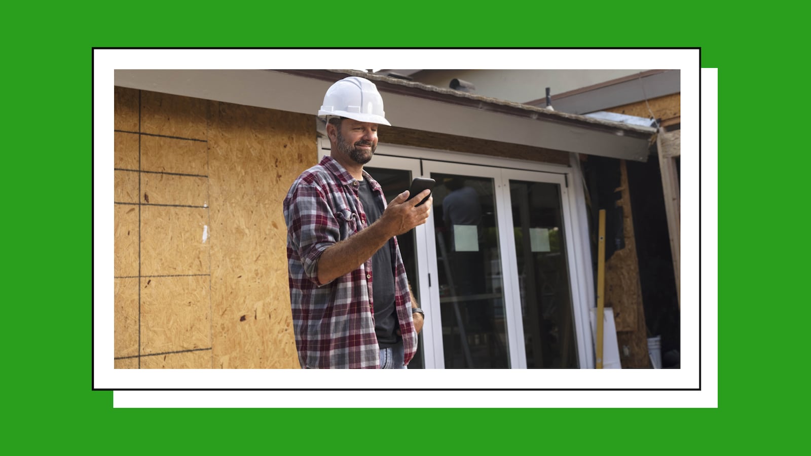 A construction worker wearing a white helmet looking at his phone. Behind him is a house under construction.