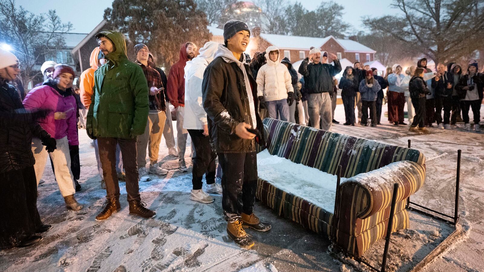 A crowd gathers in the snow. One boy stands next to a couch poised at the top of the hill.