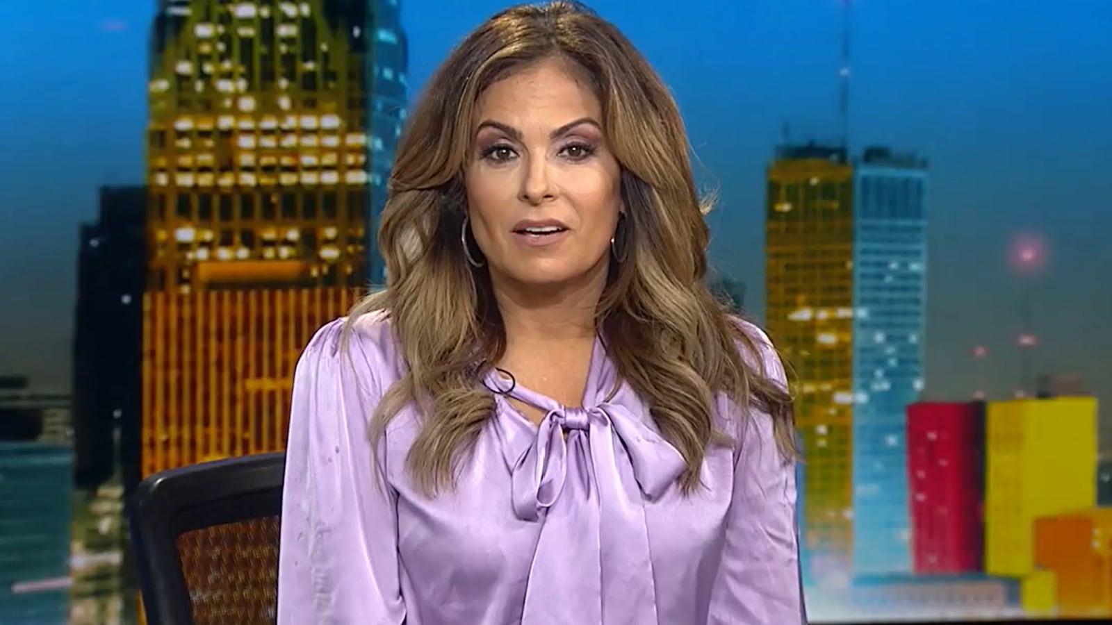 A woman in a purple blouse is sat at a TV anchor's desk, about to introduce a news story.