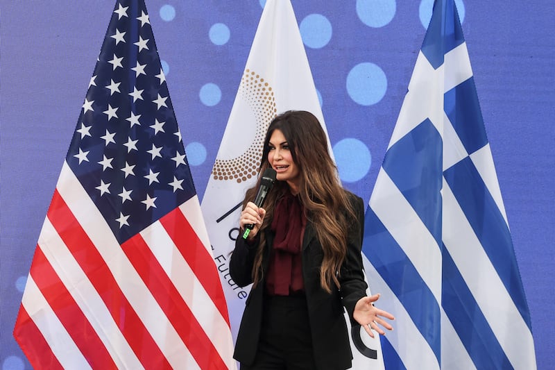 Kimberly Guilfoyle standing between the flags of the U.S. and Greece