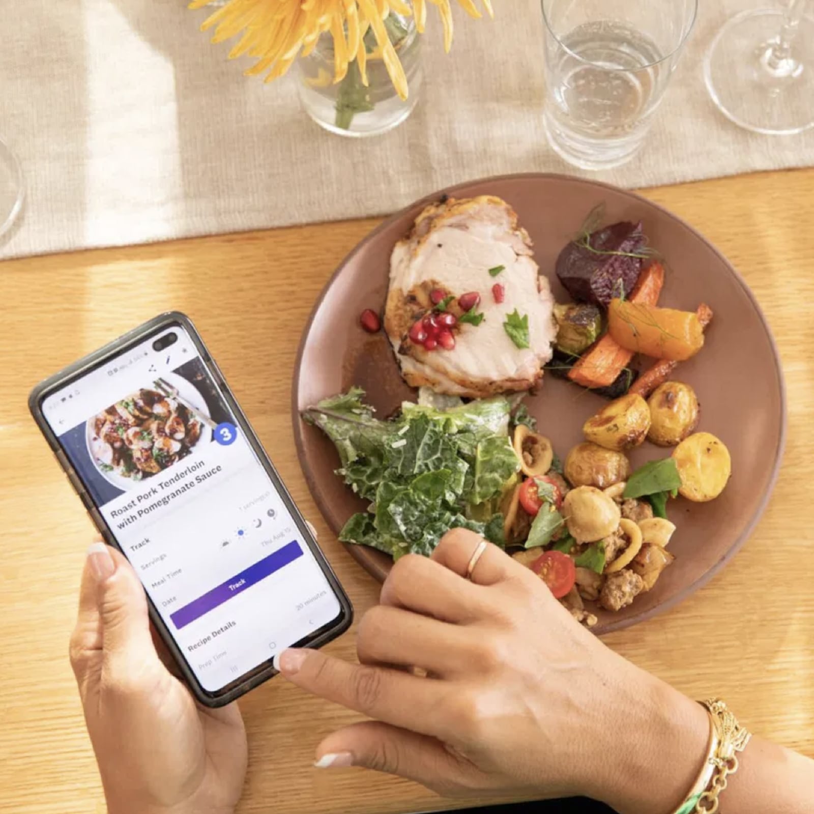 Person using the Weight Watchers app on a smartphone while eating a balanced meal with roasted chicken, vegetables, and salad at a dining table.