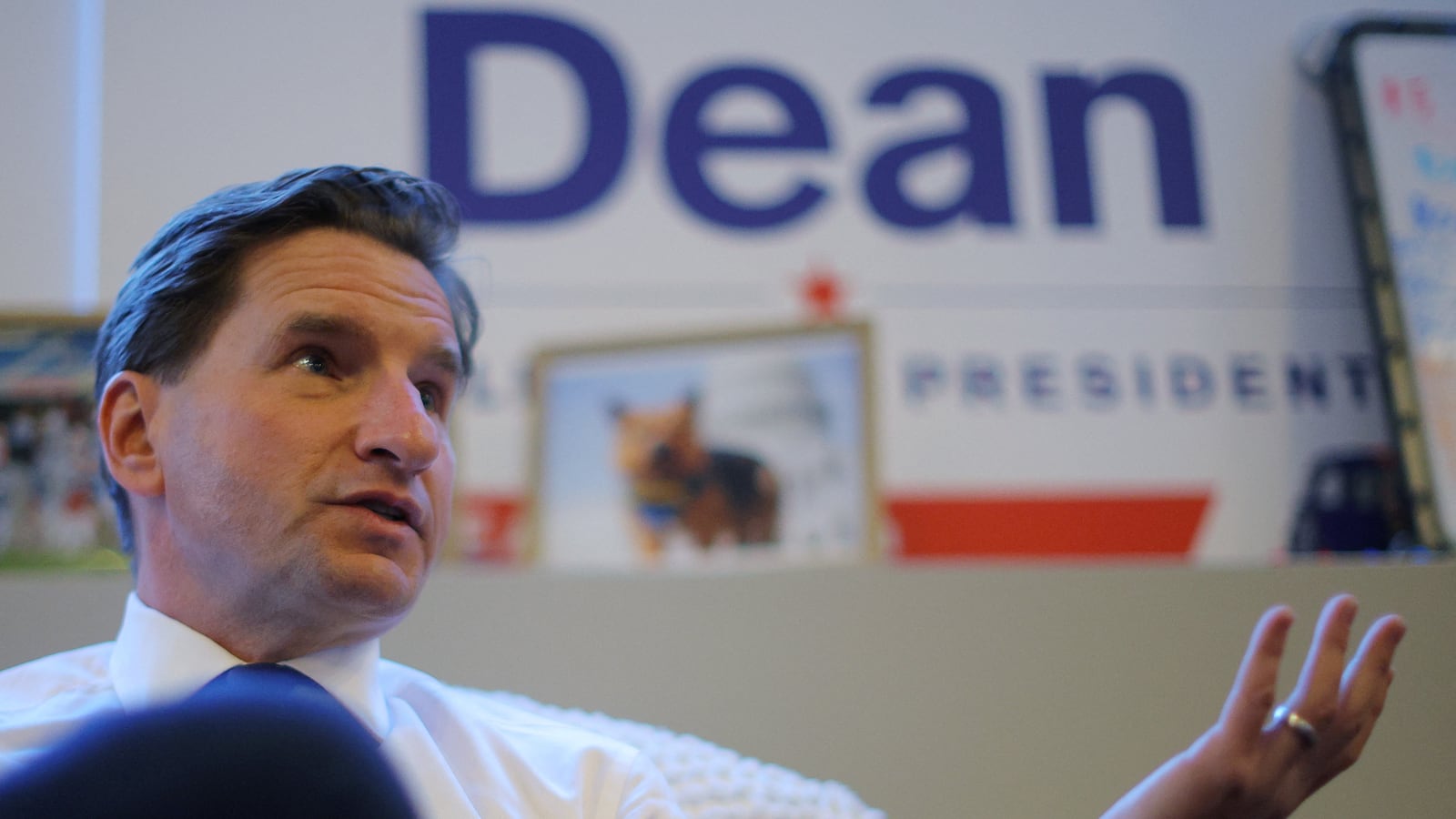 Dean Phillips, wearing a shirt and tie, speaks during an interview, with a “Dean For President” sign in the background.
