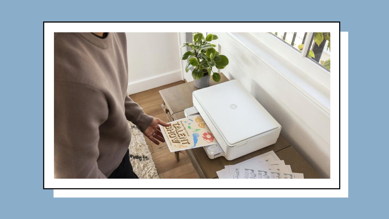 Person using an HP printer to print a colorful 'Talent Show' flyer, with a plant and printed sheets nearby on a wooden table next to a window.