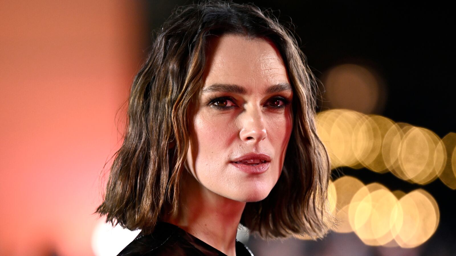 Keira Knightley looks on at night with lights behind her.