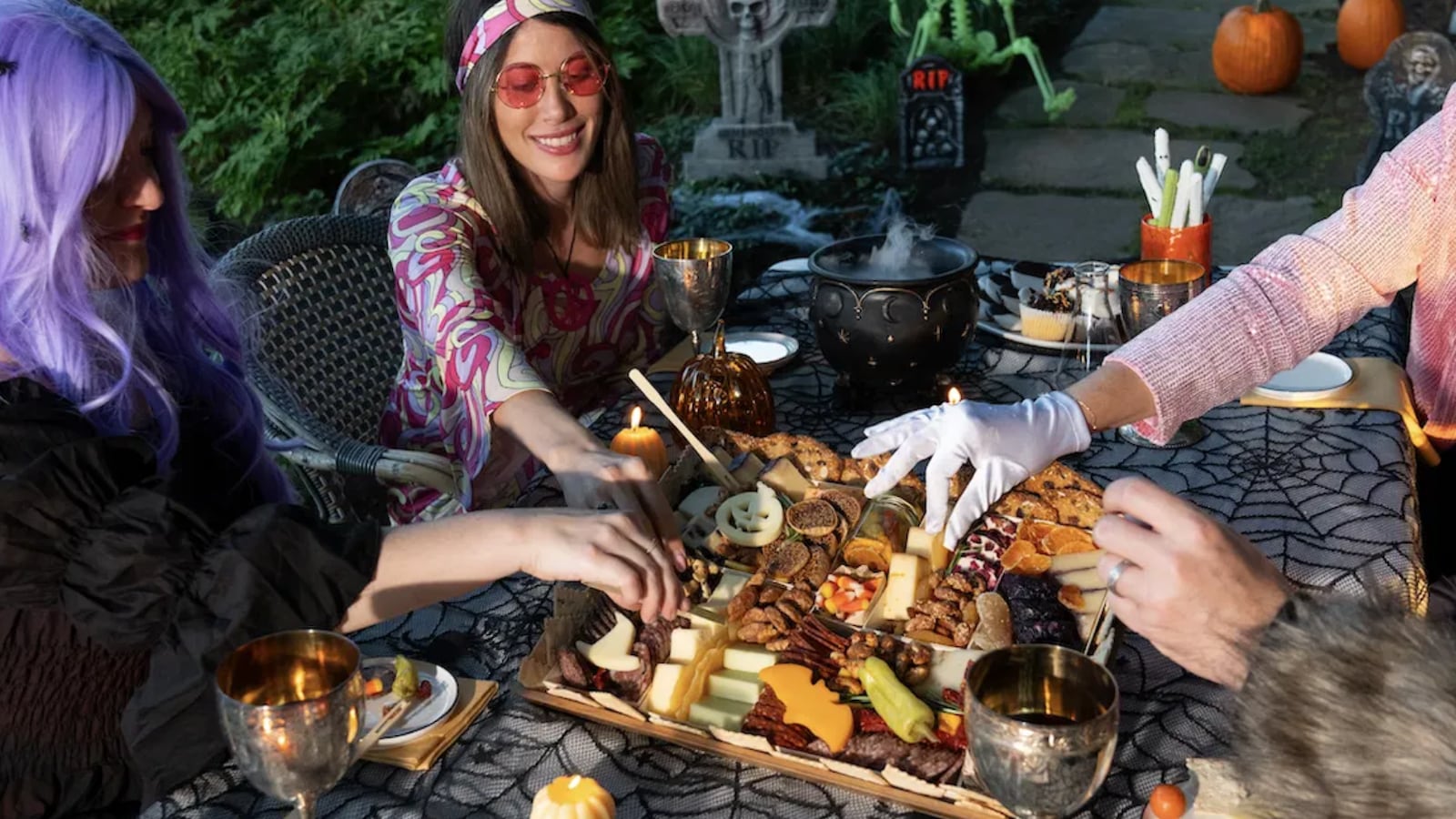 Group of friends enjoying a Halloween-themed charcuterie board at a festive outdoor gathering, surrounded by decorations like pumpkins and a spooky cauldron, featuring a variety of cheeses, meats, and treats