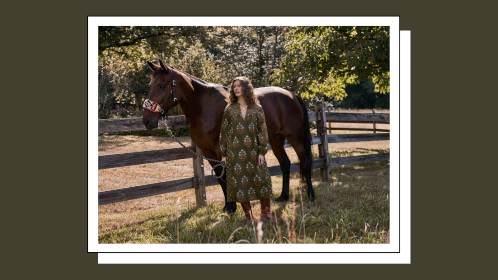 A woman in an olive floral midi dress and brown knee-high boots stands beside a brown horse at a wooden fence in a sunny pasture.