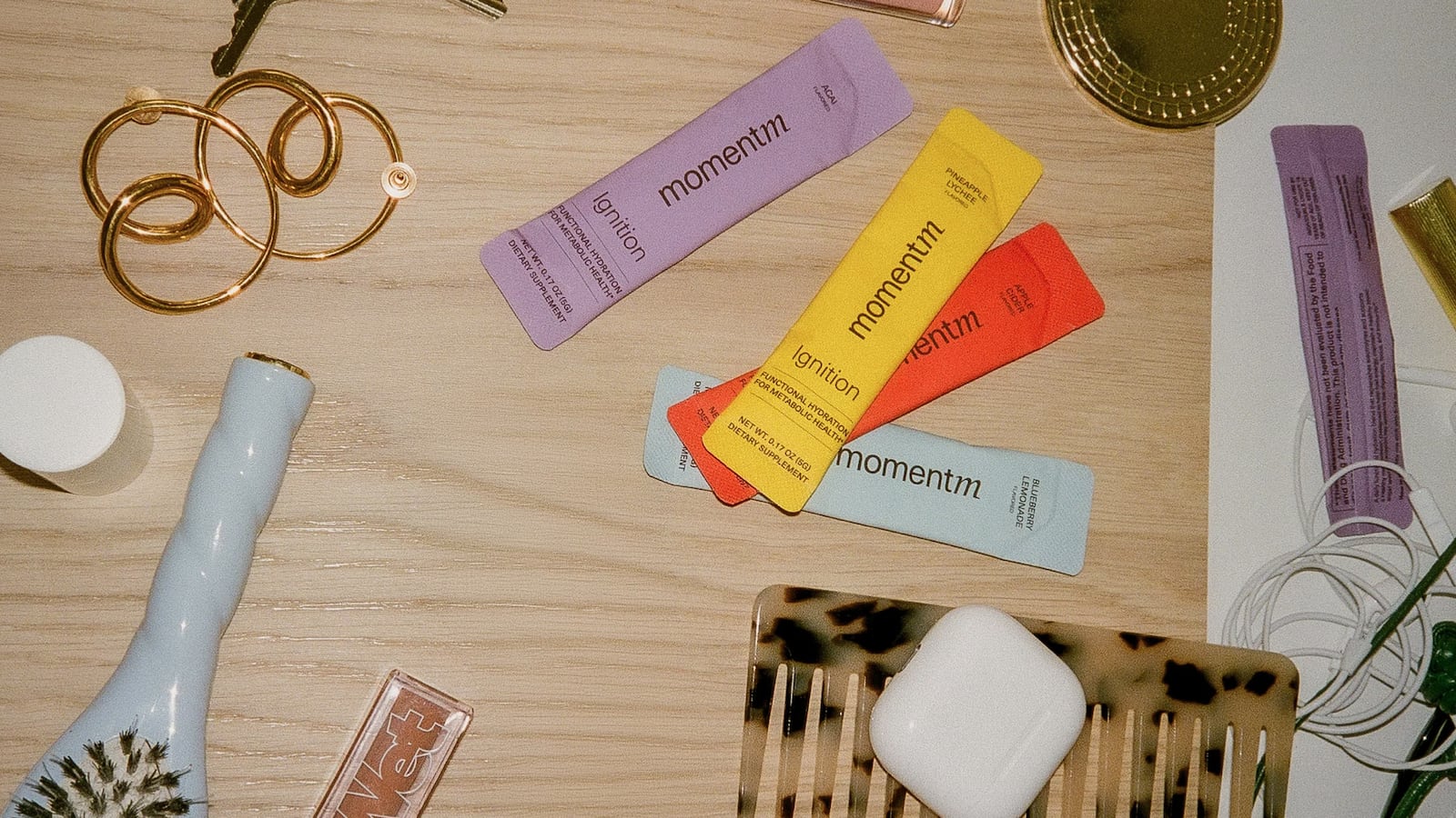 Four momentm Ignition hydration packets in açai, pineapple lychee, apple cider, and blueberry lemonade flavors, arranged on a wooden surface alongside gold jewelry, a tortoiseshell comb, and wireless earbuds.