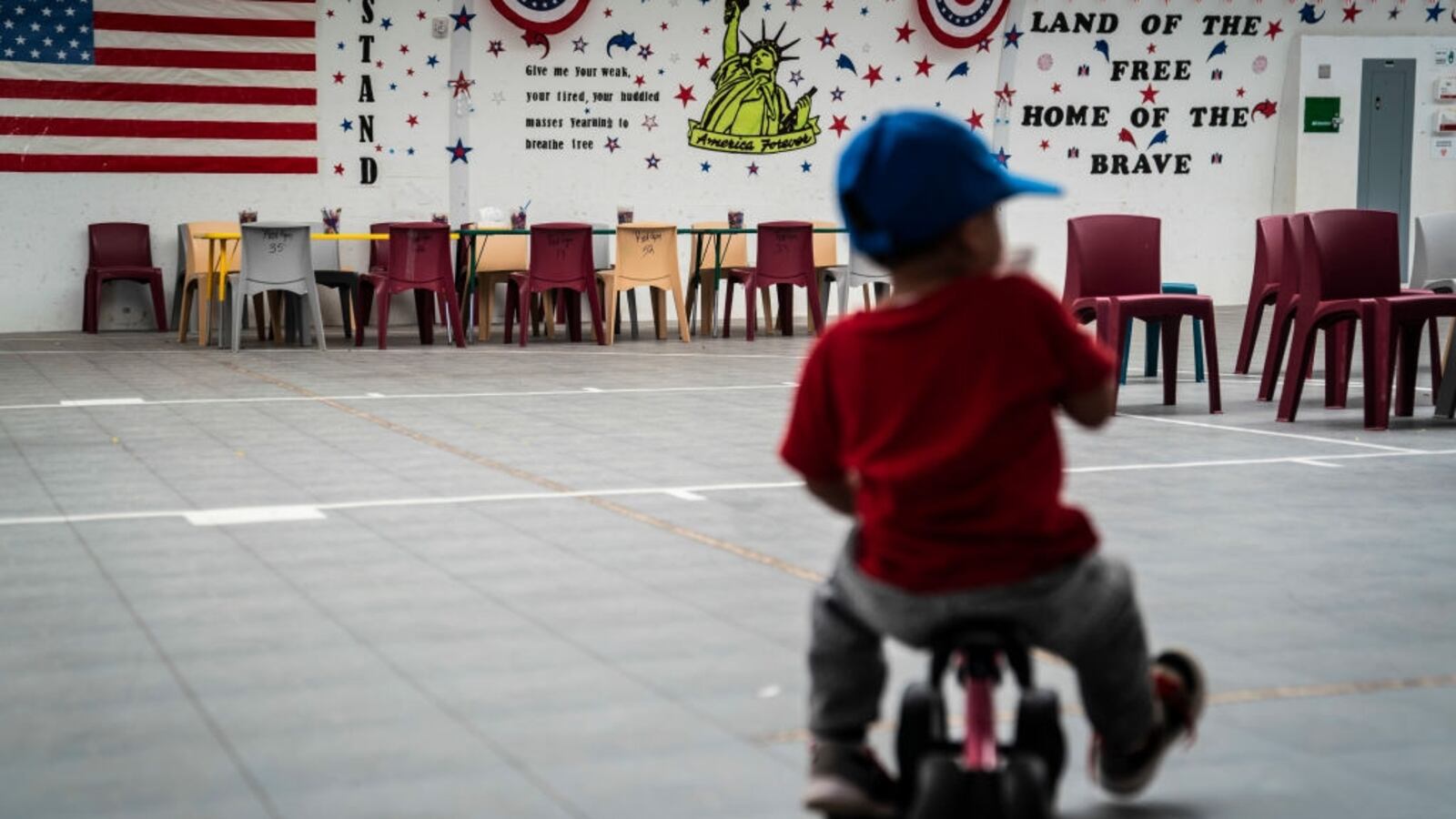 An immigrant child plays in front of patriotic phrases and symbols covering the walls in the gymnasium of an ICE facility.