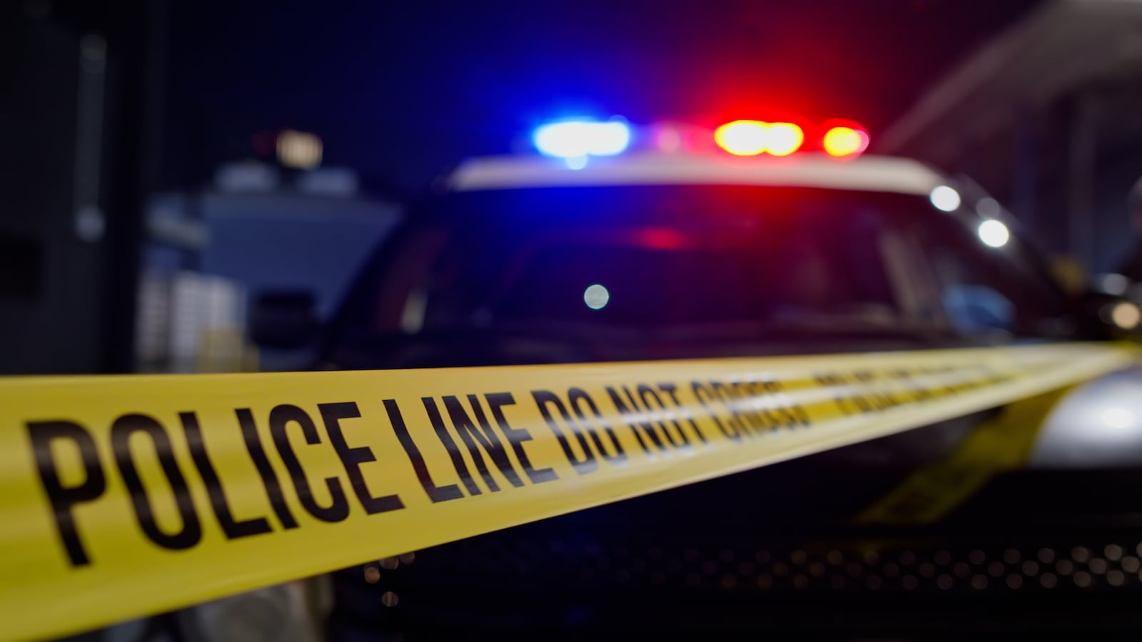A police crime scene at night with yellow police tape in the foreground reading 'POLICE LINE DO NOT CROSS' and emergency vehicles with flashing red and blue lights in the background. The scene is illuminated by the colorful emergency lighting creating a dramatic atmosphere typical of law enforcement investigations and crime scene documentation.