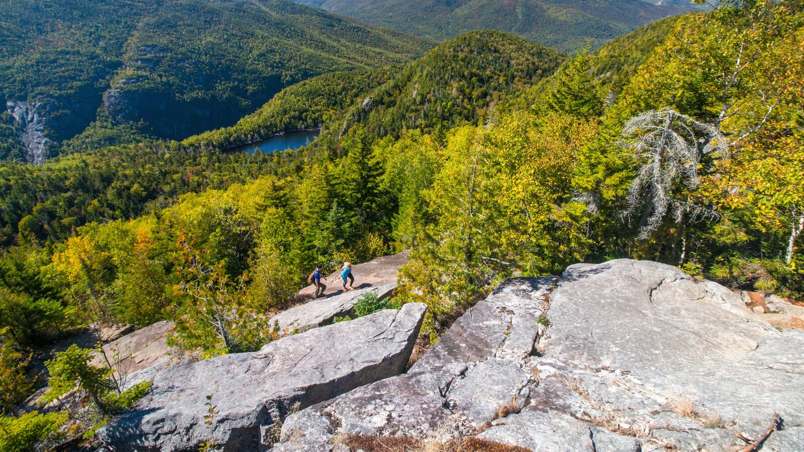 Young couple clambering up rocky slope overlooking New England's Adirondack Park in autumn, upstate New York, USA.