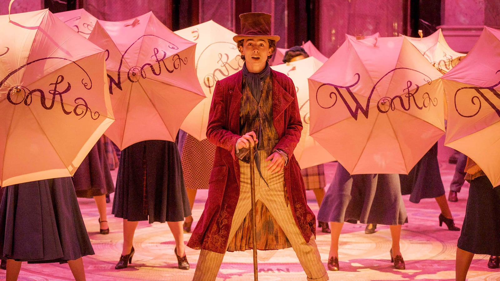 Timothée Chalamet as Willy Wonka stands in the middle of a pink room, in front of several people holding umbrellas that say “Wonka” on them.