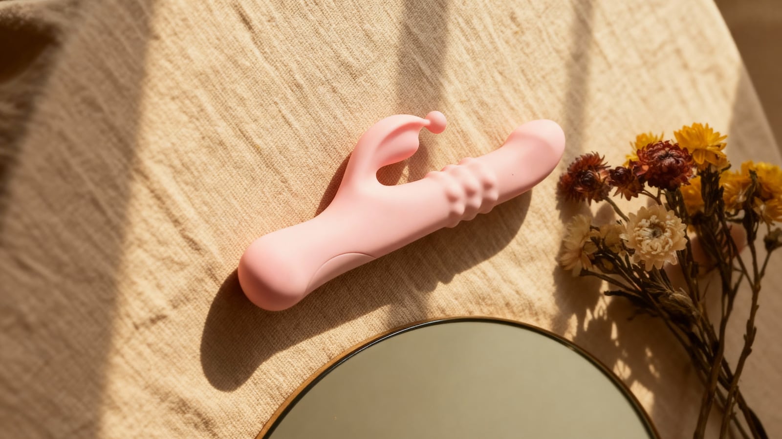Pink rabbit vibrator resting on beige fabric beside dried flowers, showcasing the Thrum’s curved clitoral arm and textured shaft.