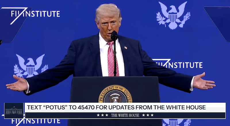 Donald Trump stands on stage at a lecture with his arms stretched out.