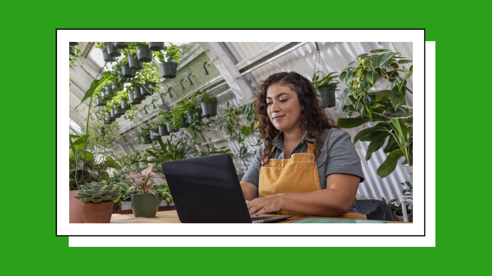 A woman working on a laptop in a building with plants all around her. She is dressed as a gardener.