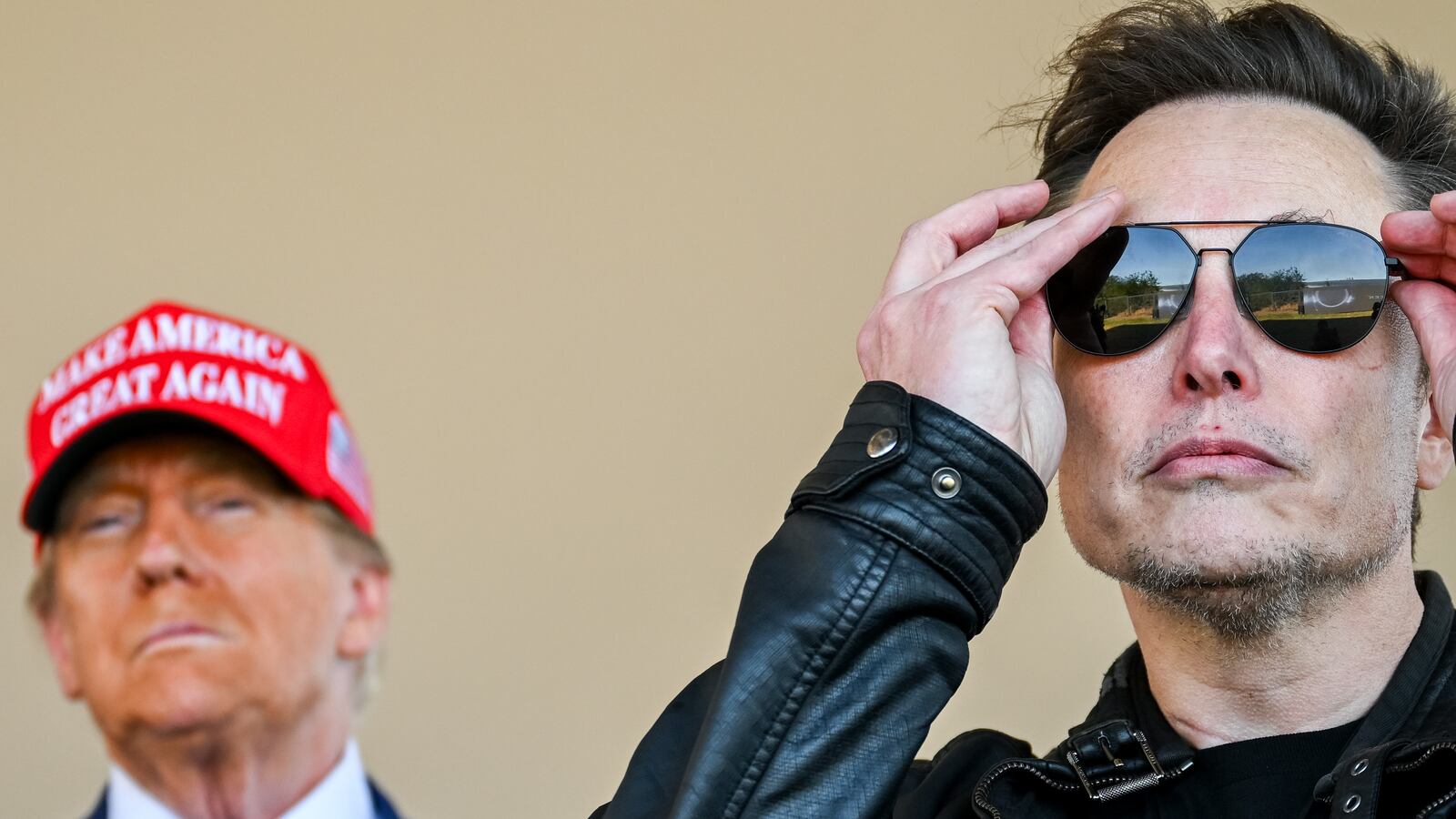 Musk puts on sunglasses with Donald Trump behind him in a MAGA hat