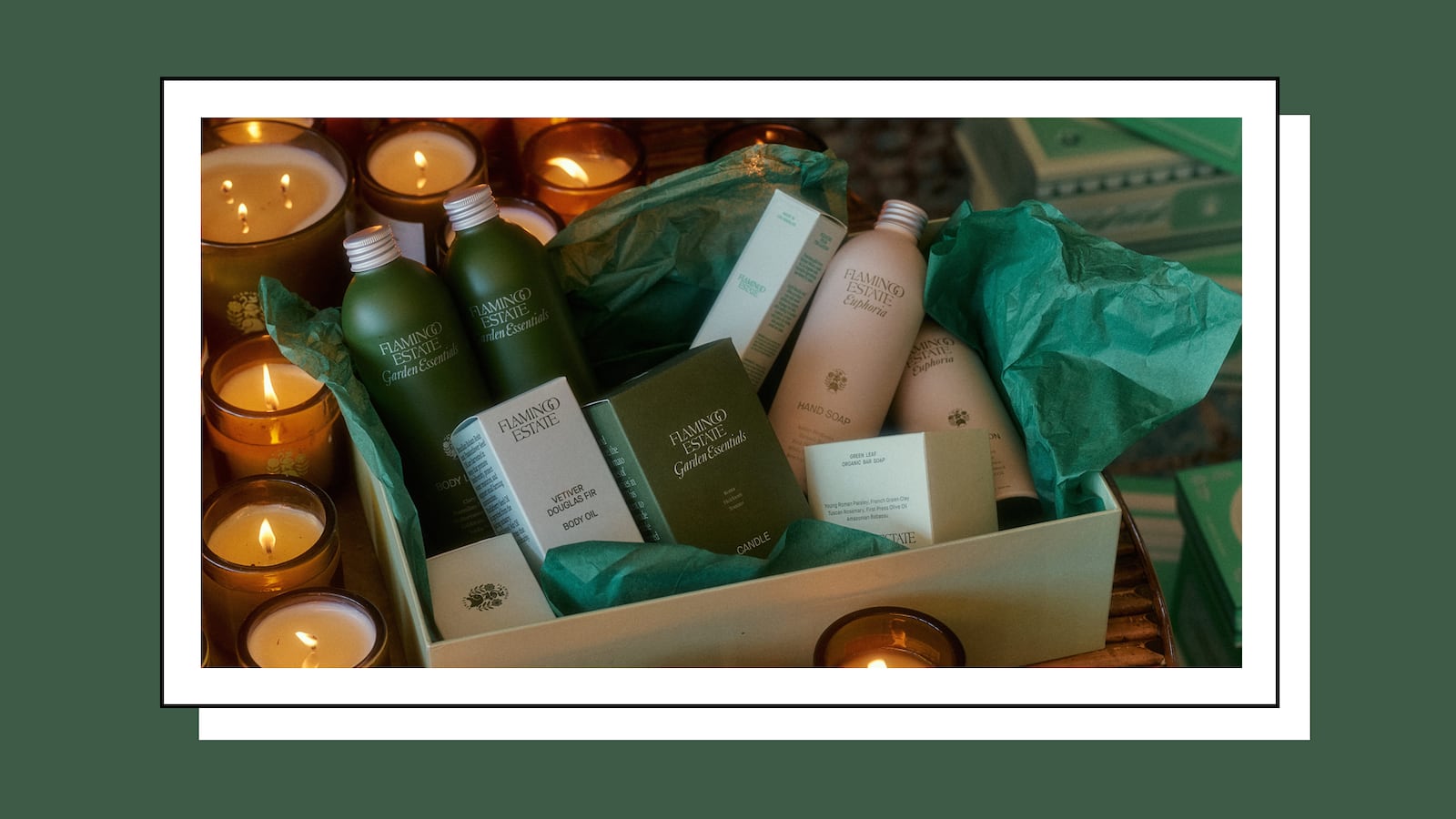 A curated gift box from Flamingo Estate, featuring a selection of luxury body care products, including hand soap, body oil, and a candle, elegantly arranged with green tissue paper. The box is surrounded by softly glowing candles, creating a warm, inviting ambiance.
