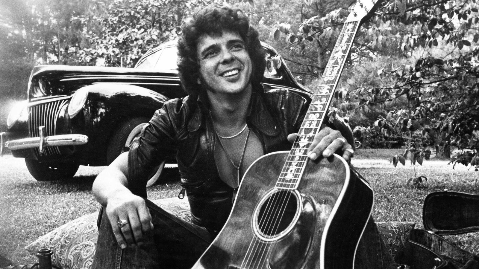 Songwriter and guitarist Bobby Whitlock poses for a portrait in front of an old car next to a bottle of Dom Perignon champagne holding an acoustic guitar in circa 1975.