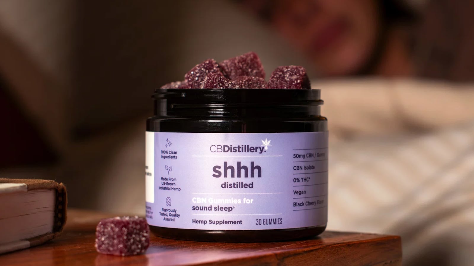 An open jar of CBDistillery Sound Sleep CBN gummies on a wooden nightstand beside a book, filled with sugar-coated black cherry gummies, with a softly blurred bedroom scene in the background