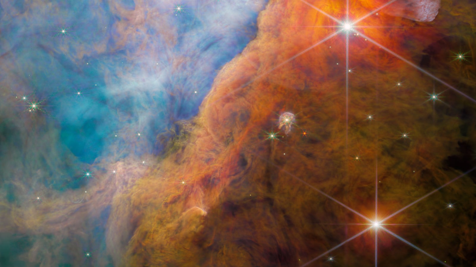 A nebula made of many layers of cloudy, colourful material. The top-left side of the image is brightly lit, filled with wispy, thin material in pale shades of pink and blue. A thick bar of denser, cloudier material crosses diagonally at the bottom right.