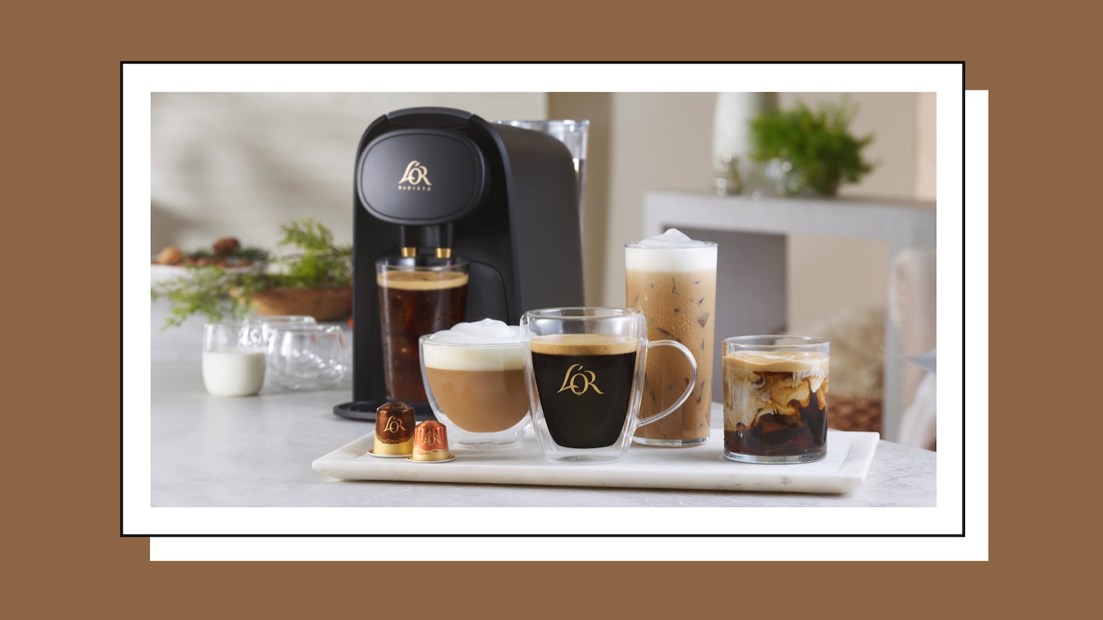 A stylish L'OR BARISTA coffee and espresso machine sits on a kitchen countertop, surrounded by various coffee drinks in elegant glassware. The drinks include a cappuccino, iced coffee, and espresso, showcasing different layers of milk and coffee. Two coffee capsules are displayed on a white serving tray in front of the machine, and a small milk jug is placed nearby.