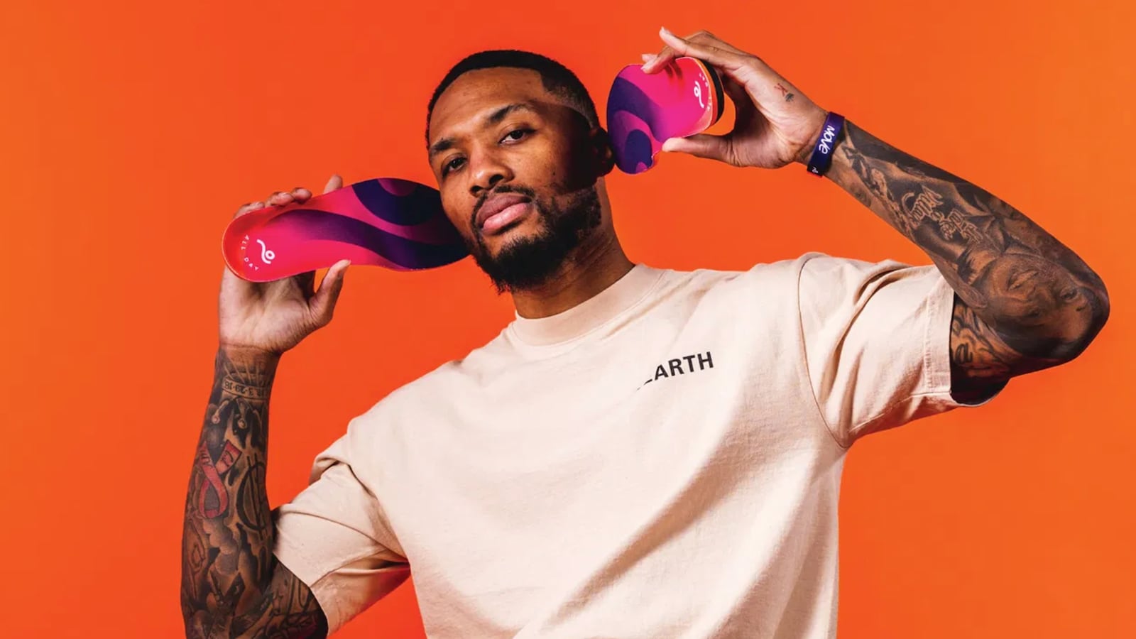 Damian Lillard standing in front of an orange background while holding two Move Insoles up to his ears.