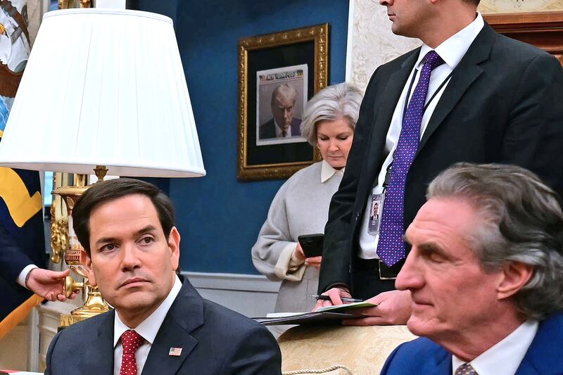 A front page of The New York Post featuring former US President Donald Trump's 2023 mugshot, taken at Fulton County Jail in Georgia, hangs on a wall outside the Oval Office. In the foreground Marco Rubio, Susie Wiles and Doug Burgum can be seen