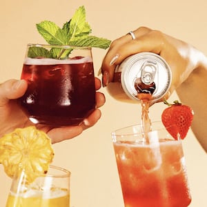 Two hands clink glasses of nonalcoholic cocktails garnished with mint, strawberry, and pineapple while a third hand pours a canned drink into a glass, set against a warm tan backdrop.