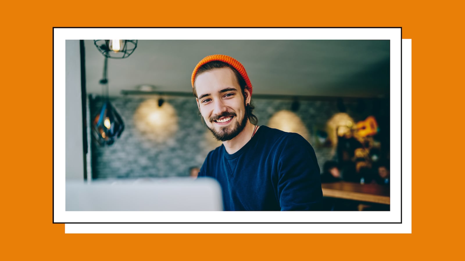 Smiling young man wearing an orange beanie and dark sweater works on a laptop in a cozy café setting, with warm pendant lights and a blurred background behind him.