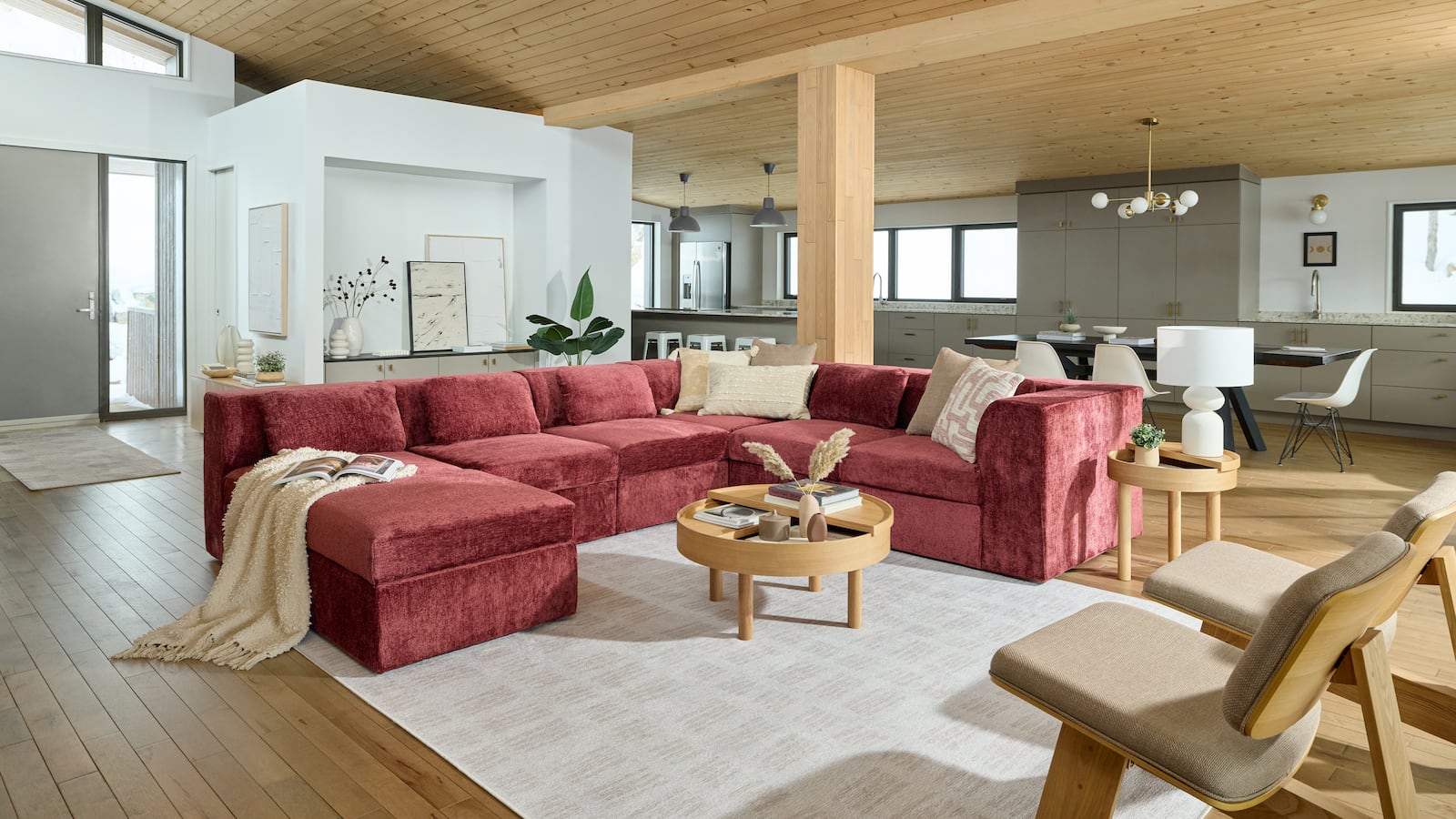 The Cozey Atmosphere couch in a dusty pink chenille sits at the center of a sun-drenched, modern room with wood and ivory touches. Light wood floors, light wood ceiling, a large ivory rug and a small, circular, light wood table with artful bric-a-brac. Ivory pillows and blankets are draped on the U-shaped couch.