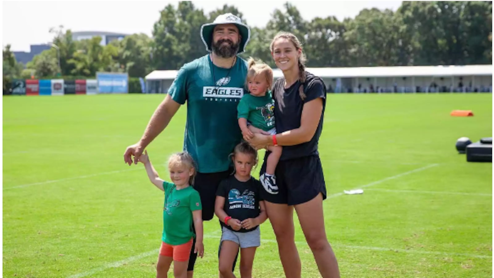 Jason and Kylie Kelce stand on a sports field with their three daughters.