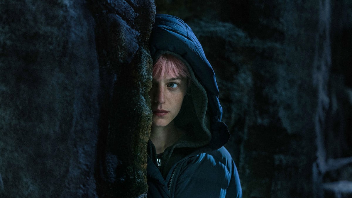Emma Corrin peering out behind a wall in a still from ‘Murder at the End of the World’