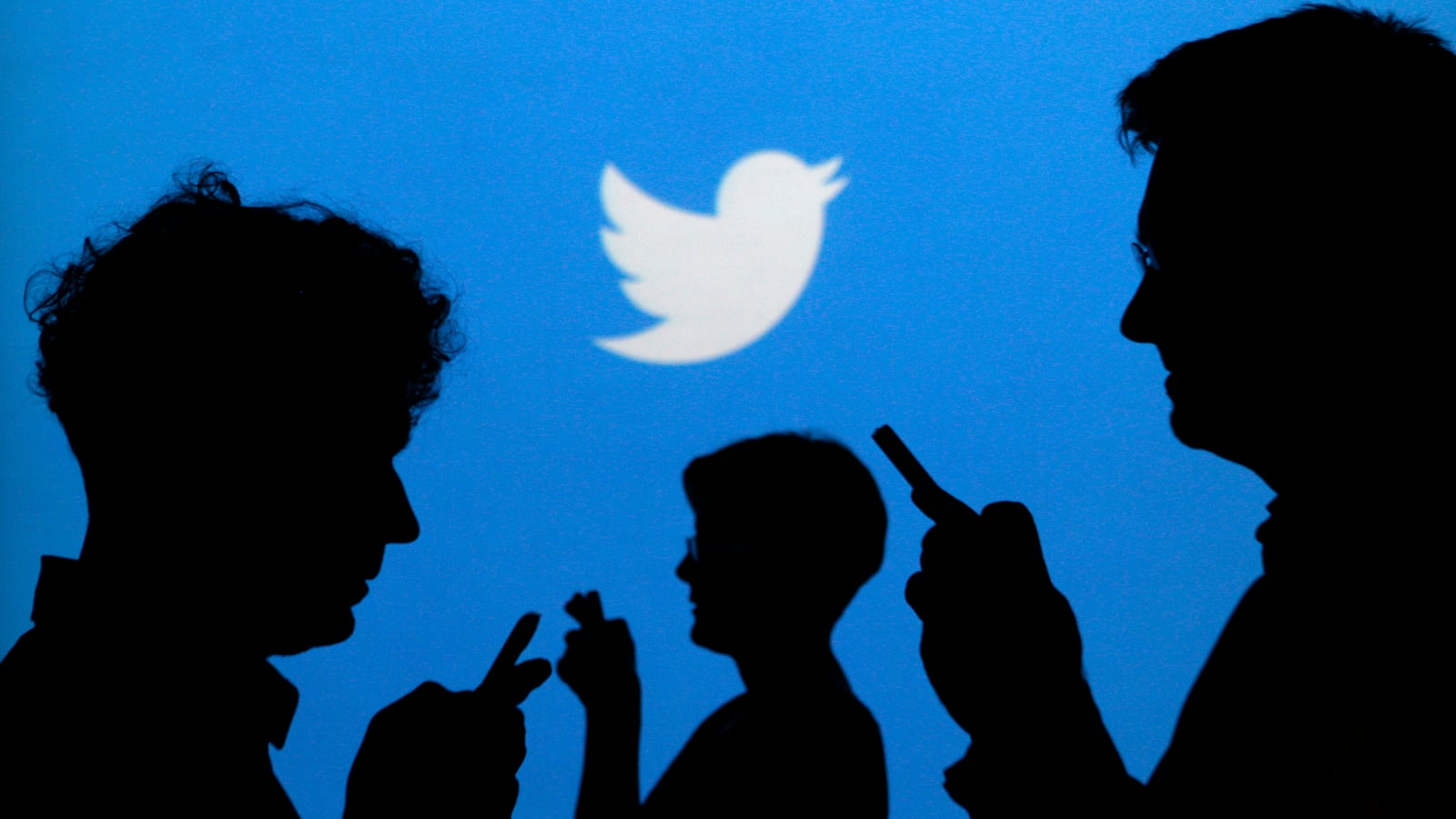 People holding mobile phones are silhouetted against a backdrop projected with the Twitter logo.