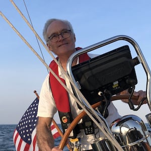Man at the helm of a sail boat with the American flag behind him.